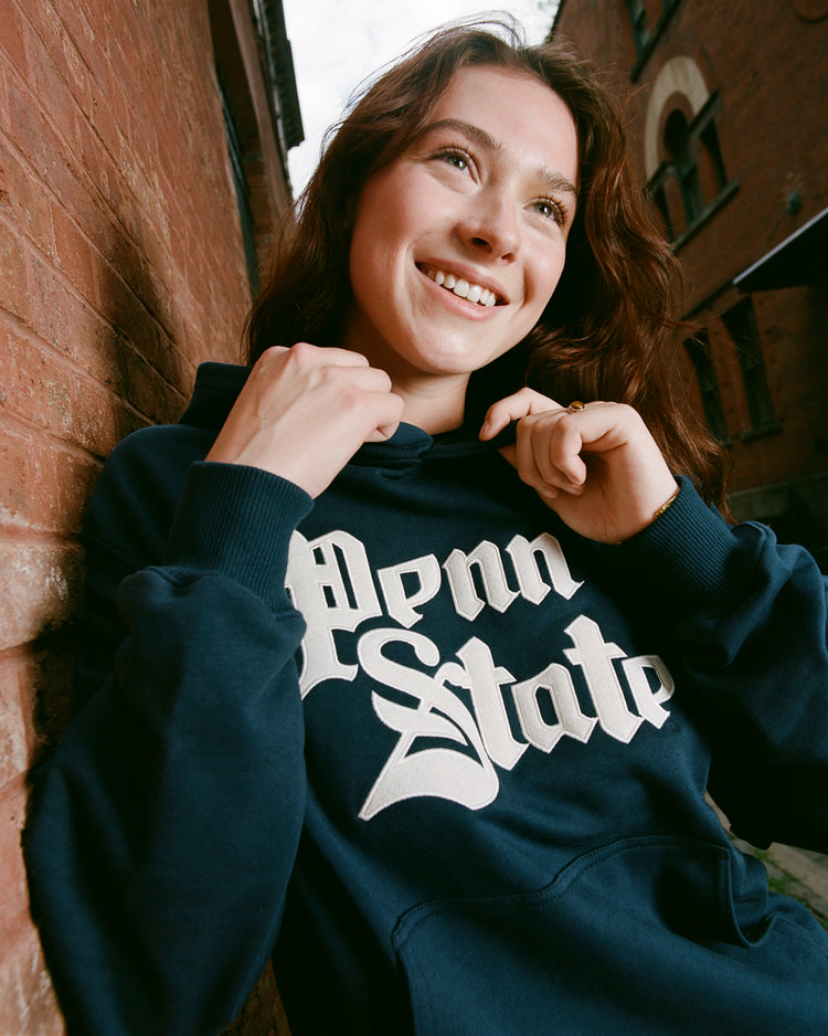 A young woman stands outside by a brick wall, wearing the Ebbets Field PENN STATE HOODIE NAVY with Penn State in bold white letters on the front.