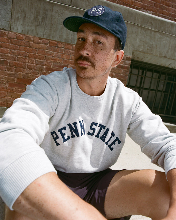 A man wearing an EFF Penn State University 1963 Authentic Wool Ballcap and a light gray PENN STATE sweatshirt with a vintage logo sits on concrete steps by a red brick wall, looking at the camera with a neutral expression.