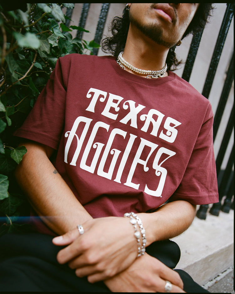 A person wearing the Ebbets Field TEXAS A&M SS TEE MAROON sits near greenery and a metal fence, accessorized with silver jewelry and a pearl necklace, their face partially out of frame.