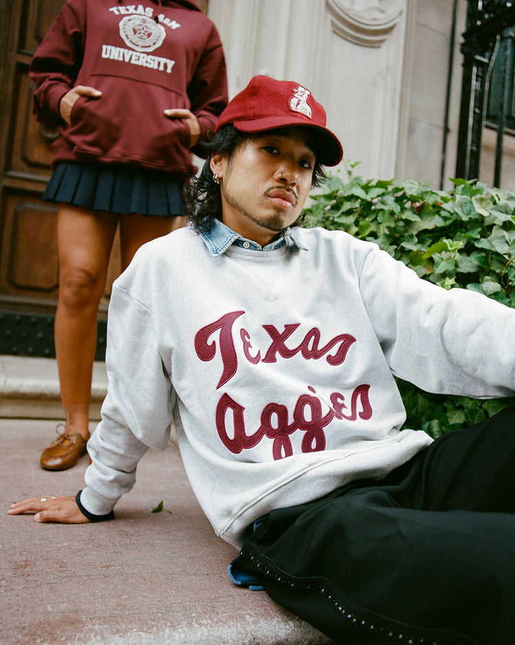 Someone in an Ebbets Field TEXAS A&M CREWNECK SS H-GRY sits on stone steps with a red cap, while another person in a similar sweatshirt and pleated skirt stands behind them near a wooden door and greenery.