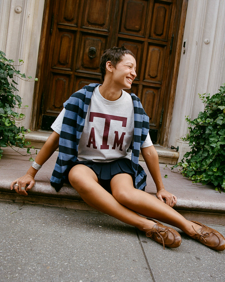 A person sits on stone steps by a wooden door, smiling. They wear the Ebbets Field TEXAS A&M SS TEE H-GRY, with a striped sweater over their shoulders, a pleated skirt, and brown shoes. Green plants line the entryway.