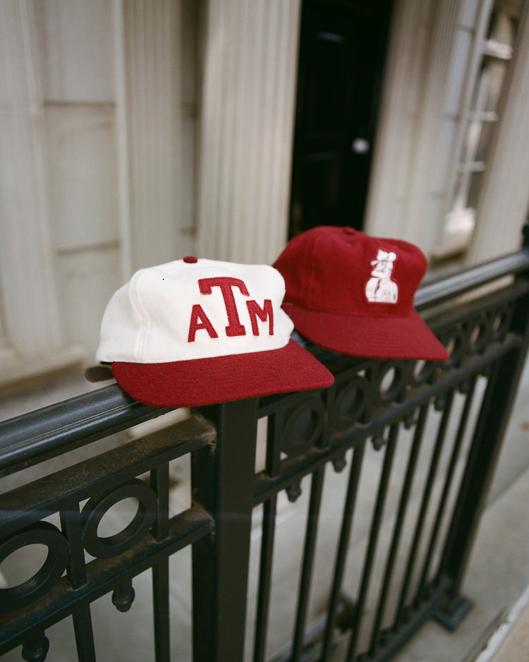Two EFF Texas A&M Aggies Collegiate Wool Ballcaps, one white with a maroon logo and one maroon with a white graphic, sit on a black iron fence. Tall white columns and windows appear in the background.