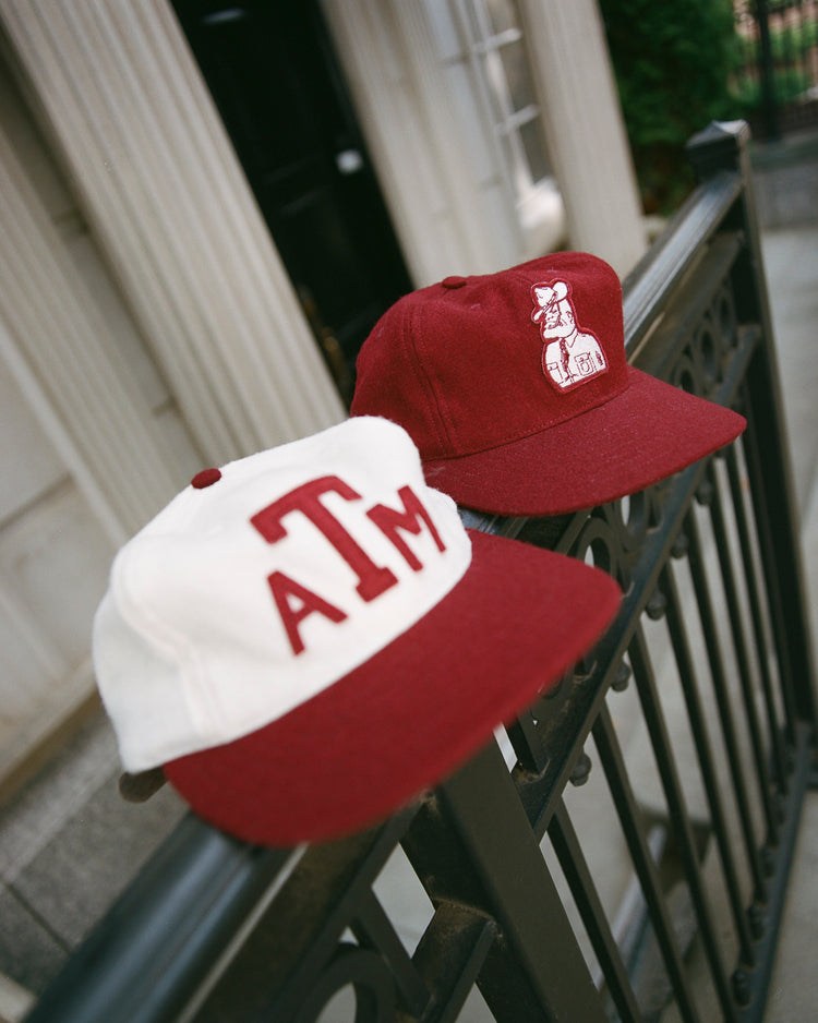 Two maroon and white EFF Texas A&M Aggies Mascot Collegiate Wool Ballcaps rest on a black wrought iron fence, with a building entrance visible in the background.