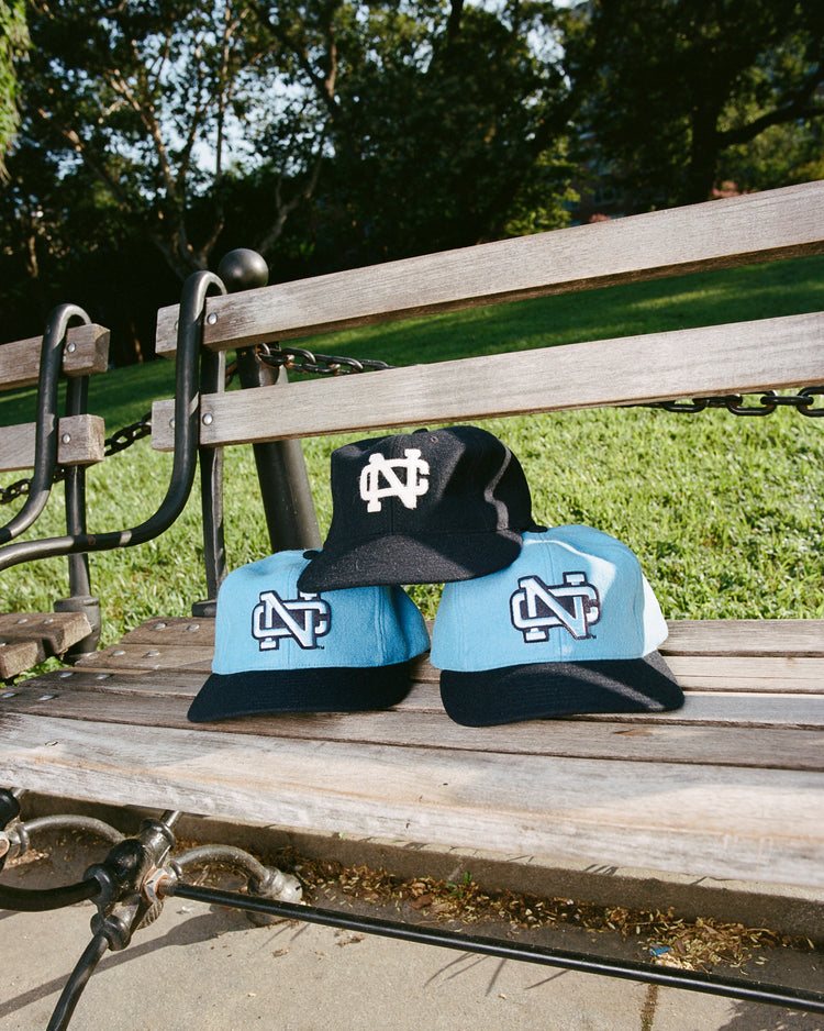 Three EFF University of North Carolina 1983 Vintage Ballcaps—two light blue with dark brims and embroidered logos, one black wool with a white logo—sit on a wooden park bench, set against grass and trees.