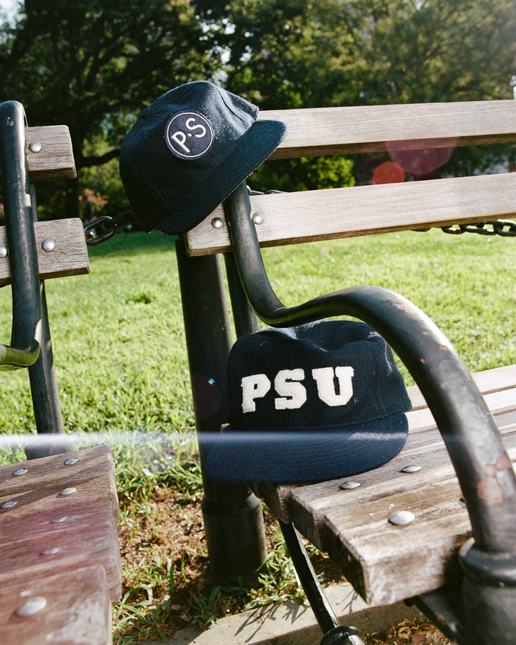 Two navy blue EFF caps sit on a wooden park bench. One is the Penn State University 1963 Authentic Wool Ballcap with a vintage Nittany Lions logo; the other features large white PSU letters. Green grass and trees are in the background.