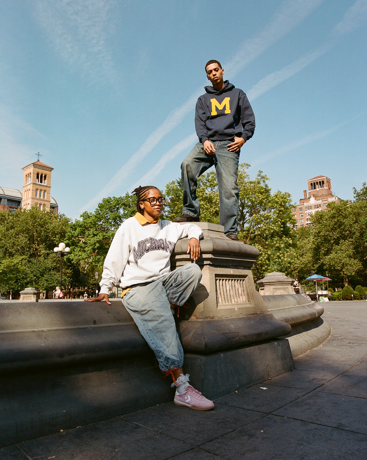 Two people pose on a stone monument in an urban park. One wears a Michigan hoodie navy by Ebbets Field with blue jeans, while the other sits in a light sweatshirt and pink sneakers. Trees and buildings are visible behind them.