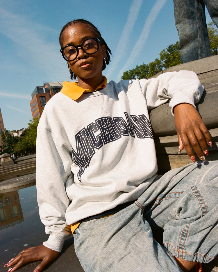 A person in large glasses, loose jeans, and the Ebbets Field MICHIGAN CREWNECK SS H-GRY sweatshirt sits by a fountain outdoors, with trees and buildings in the background under a blue sky with wispy clouds.