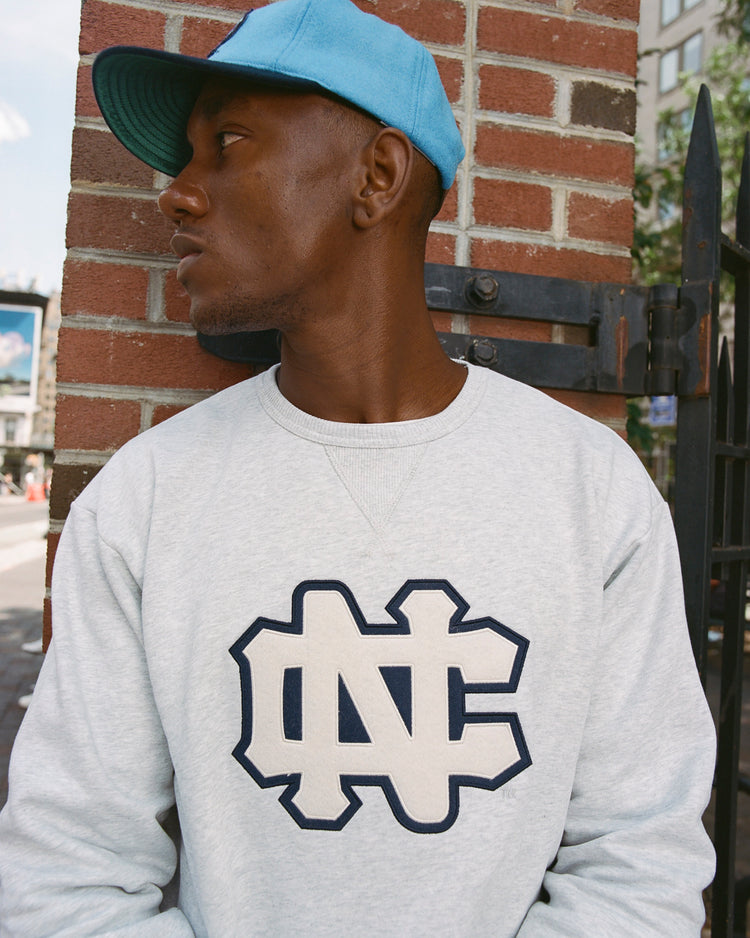 A person in an Ebbets Field UNIVERSITY OF NORTH CAROLINA CREWNECK SS H-GRY sweatshirt and a light blue cap stands outside, leaning against a brick wall and looking to the side.