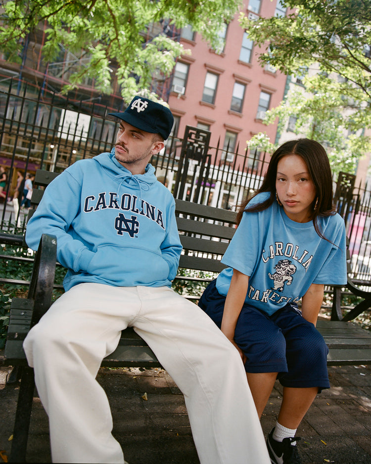 A man in a blue Carolina hoodie and an Ebbets Field Flannels University of North Carolina 1960 Vintage Ballcap sits on a bench beside a woman in a blue Tar Heels T-shirt and navy shorts, with trees and a brick building behind them.