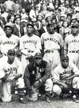 A vintage black-and-white photo of an all-Black baseball team wearing Yankees uniforms, posed together on a field with a crowd of spectators in the background.