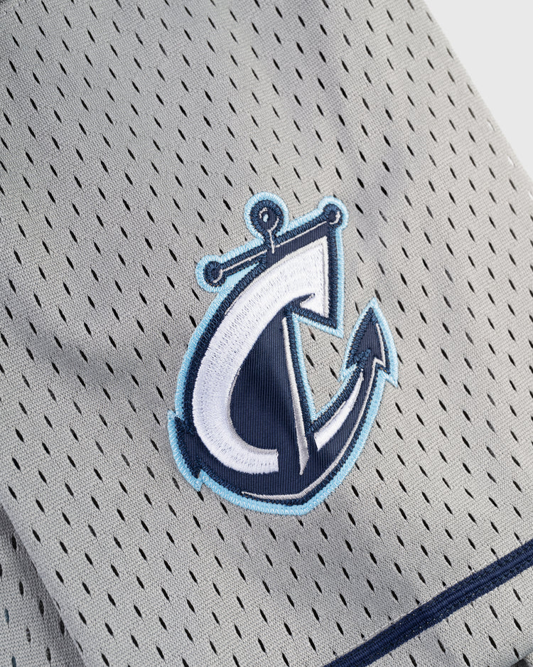 A close-up of the FRANCISCO LINDOR BUTTON UP MESH JERSEY GRY by Ebbets Field, showing gray mesh fabric with an embroidered blue and white anchor logo featuring an integrated letter C.