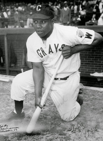A Black baseball player in a GRAYS uniform and a dark cap adjusts his sleeve on the field, with spectators visible in the blurred background.
