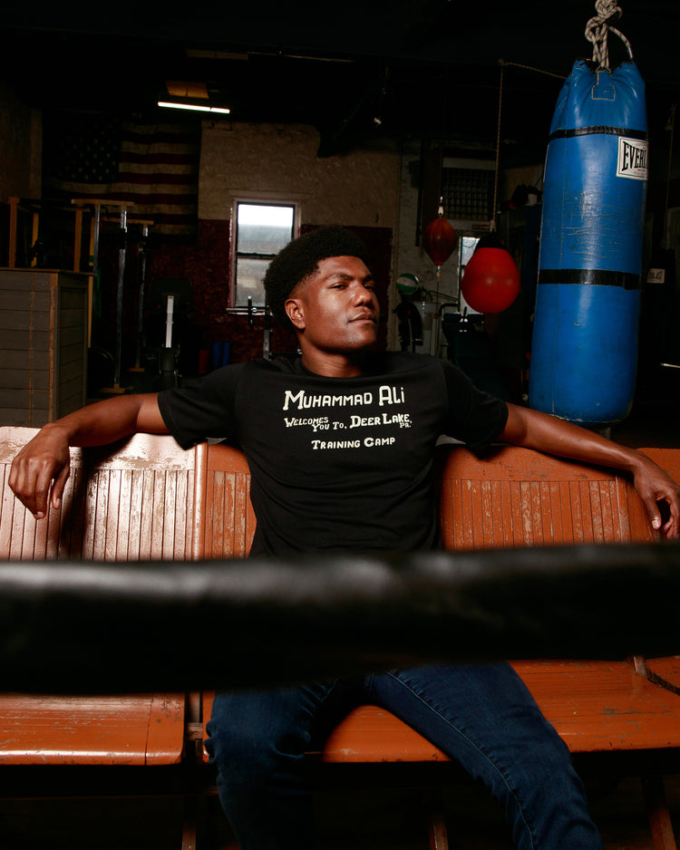 A man sits on a wooden bench in a boxing gym, wearing an Ebbets Field Muhammad Ali 'Deer Lake Training Camp' Short Sleeve Graphic Tee, with blue boxing bags and equipment in the background.