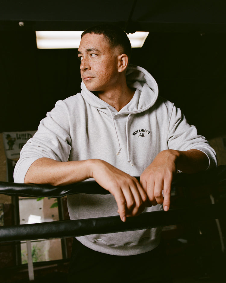 Wearing an Ebbets Field Muhammad Ali Chest Embroidered Heavyweight Cotton Terry Pullover Hoodie, a person leans on boxing ring ropes in a dimly lit gym, gazing thoughtfully to the side.