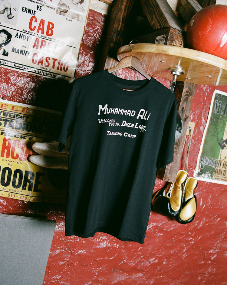 The Ebbets Field Muhammad Ali 'Deer Lake Training Camp' Short Sleeve Graphic Tee hangs on a wooden hanger, surrounded by vintage boxing posters and gloves that honor Ali and iconic bouts like the Thrilla in Manila.