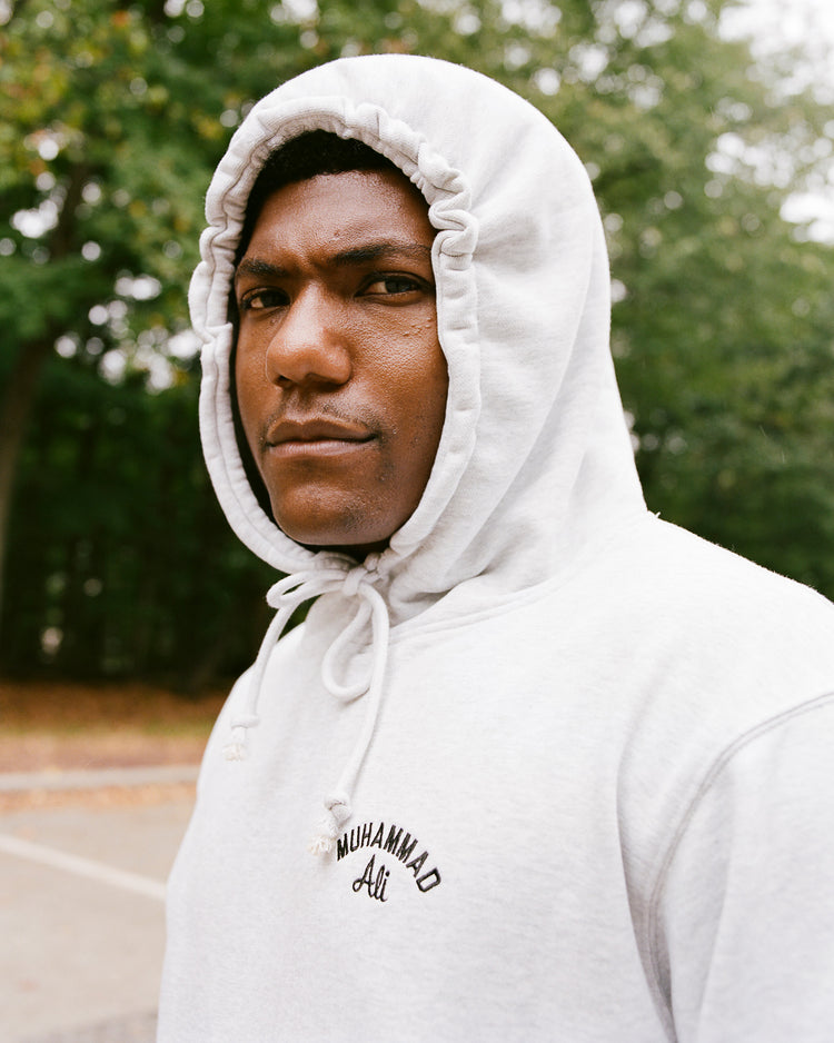 A person wears an Ebbets Field Muhammad Ali Chest Embroidered Heavyweight Cotton Terry Pullover Hoodie outdoors on a cloudy day, framed by trees and looking directly at the camera with a serious expression.