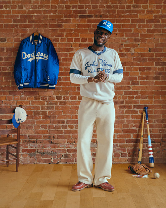 A smiling man wearing a Jackie Robinson All Stars jersey, white pants, and a blue 42 cap stands in front of a brick wall. Nearby are a Dodgers jacket, bats, gloves, and a cap on a chair.