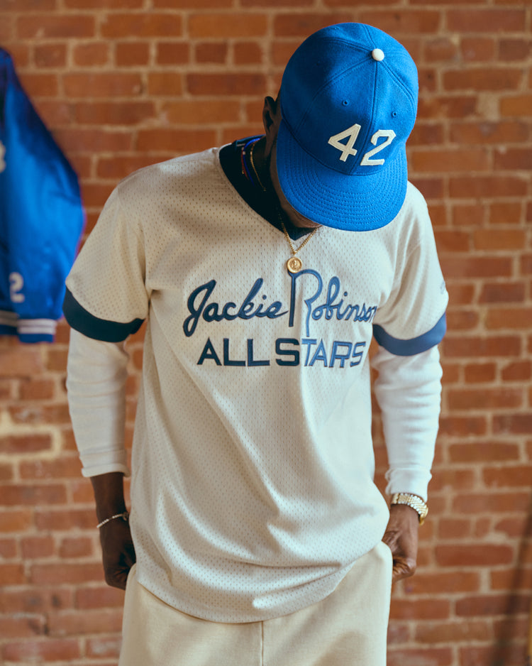 Wearing an Ebbets Field Jackie Robinson Mesh V-Neck Jersey and a blue 42 cap, a person stands against a brick wall, looking down—a tribute to baseball history and the All Stars’ enduring legacy.