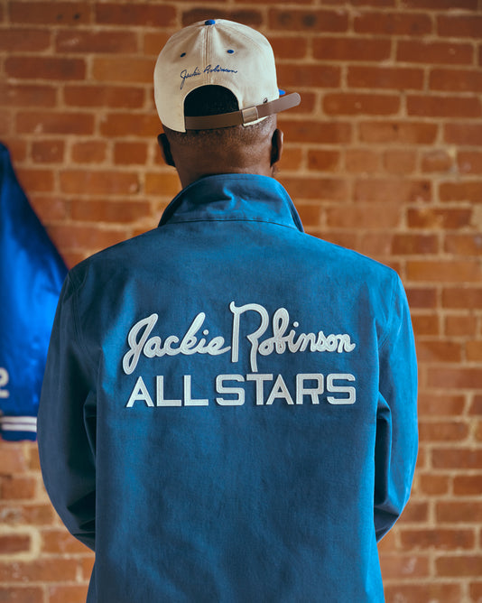 A person wearing a blue jacket with Jackie Robinson ALL STARS written on the back and a beige cap stands facing a brick wall.