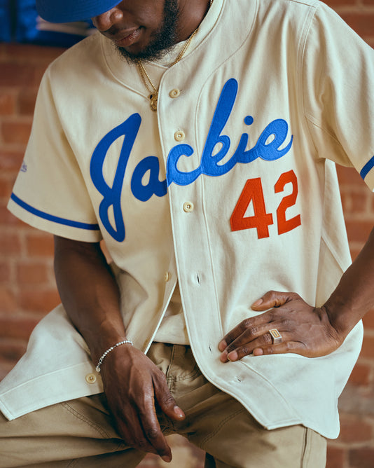 A person wearing a cream-colored baseball jersey with Jackie in blue script and the number 42 in red, paired with tan pants and gold jewelry, sits in front of a brick wall.
