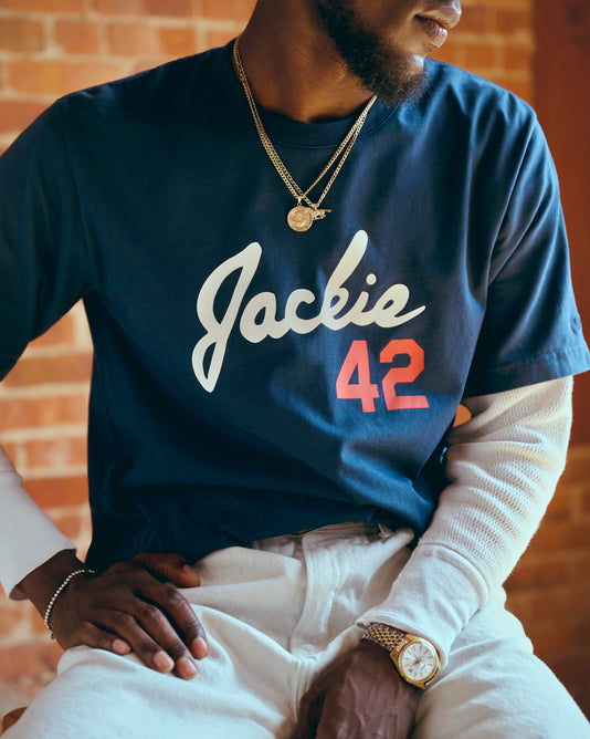 A man wearing layered shirts sits with his hands on his lap. His navy shirt has “Jackie 42” written on it in white and red, and he wears gold necklaces and a watch. The background consists of brick walls.