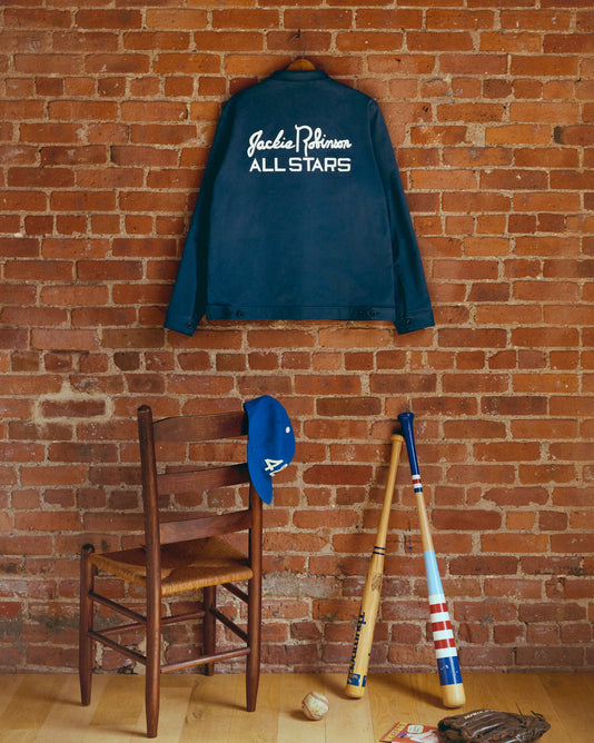 A navy Jackie Robinson All Stars jacket hangs on a brick wall above a wooden chair with a blue cap. Two baseball bats, a glove, and a baseball are on the floor in front of the chair.