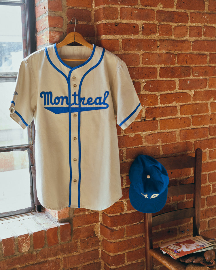 The Ebbets Field Montreal Royals Cotton Twill Baseball Jersey hangs on a wooden hanger by a brick wall, beside a matching blue cap on a chair with magazines, as sunlight pours through the window.