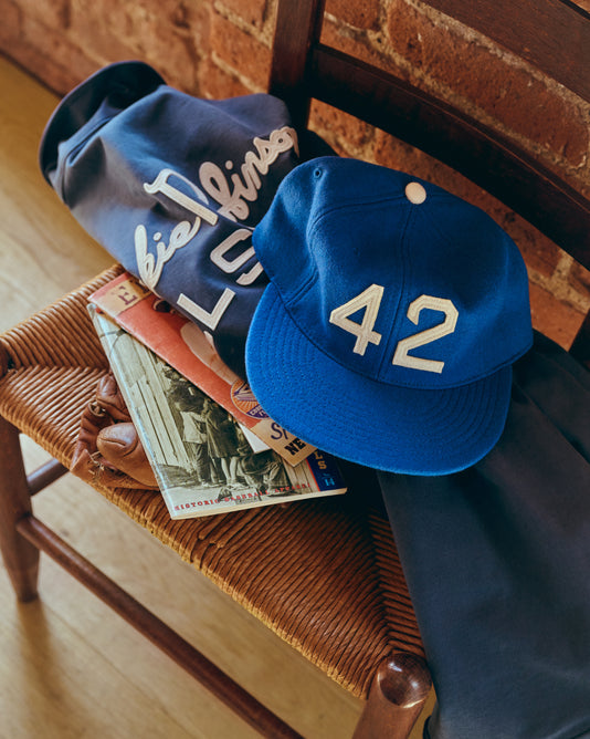 A blue baseball cap with the number 42 sits on a chair along with a blue jacket, a baseball glove, and vintage magazines featuring a photo of a baseball player. A brick wall is in the background.