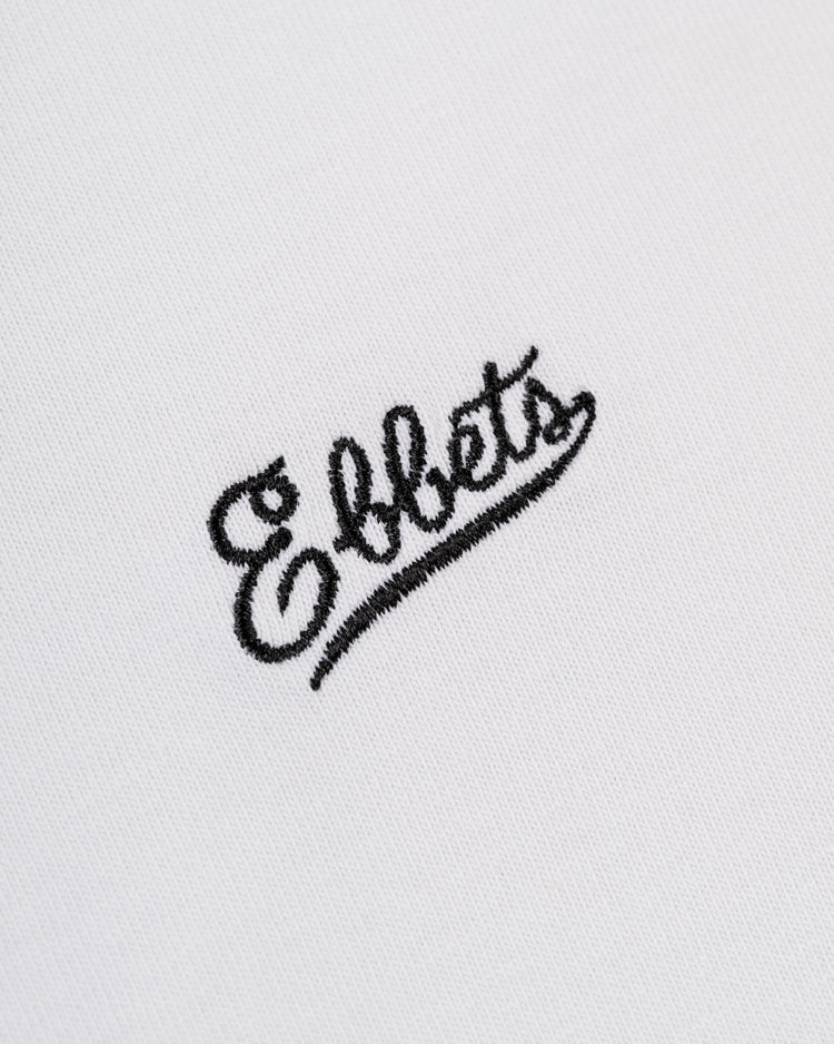 Close-up of the embroidered Ebbets Field Flannels chest logo in black cursive on the white Ebbets Chest Logo Short Sleeve Tee—a classic made from durable cotton.