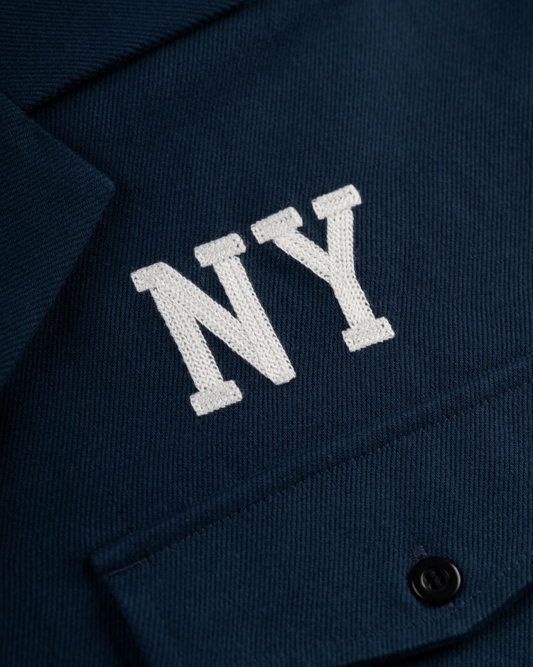 Close-up of the Ebbets Field Flannels Ebbets Cotton Linen Grounds Crew Short Sleeve Workshirt in navy, with white NY embroidery above the buttoned pocket.