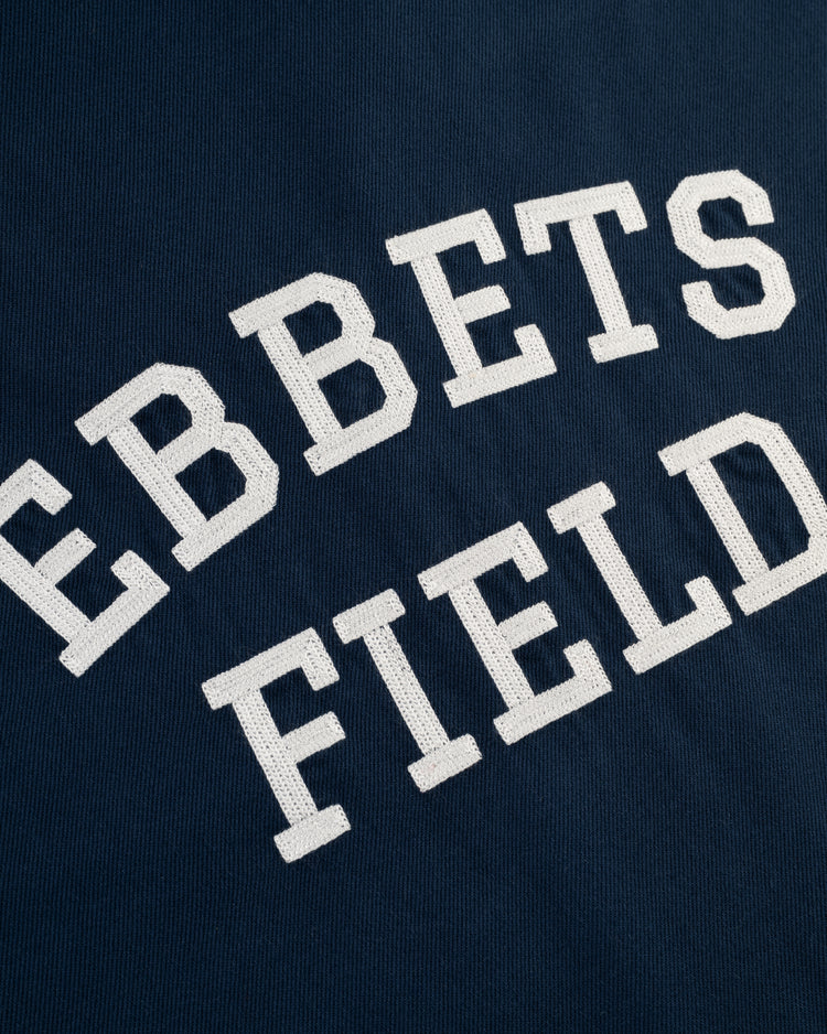 The Ebbets Cotton Linen Grounds Crew Short Sleeve Workshirt by Ebbets Field Flannels features navy blue fabric and bold, stitched white “EBBETS FIELD” letters in two curved lines, inspired by classic workwear in premium cotton linen.