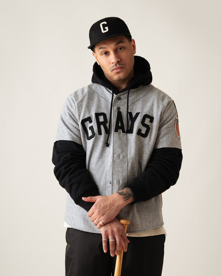 A man wears an Ebbets Field Flannels Homestead Grays Felt G Cotton Twill Ballcap, a gray Grays baseball jersey, and a black hoodie, holding a bat and looking neutrally at the camera against a plain background.