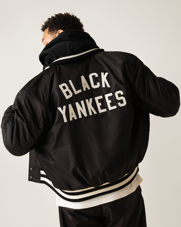 Someone faces away from the camera wearing the Ebbets Field New York Black Yankees Satin Varsity Jacket, featuring bold white BLACK YANKEES lettering and white stripes on the collar and hem, inspired by Negro Leagues Baseball Museum heritage.