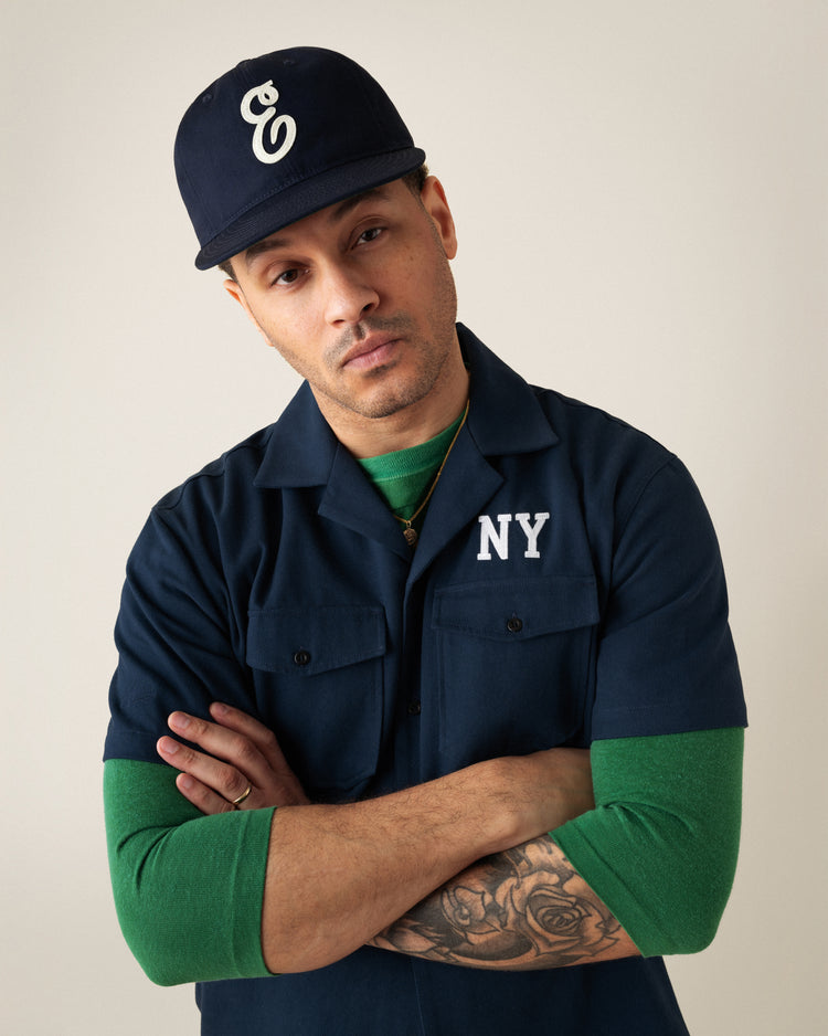 A tattooed man stands with arms crossed, wearing the Ebbets Field Flannels Ebbets Felt E Cotton Twill Ballcap and a navy NY shirt over a green long-sleeve—an effortless everyday look against a plain background.