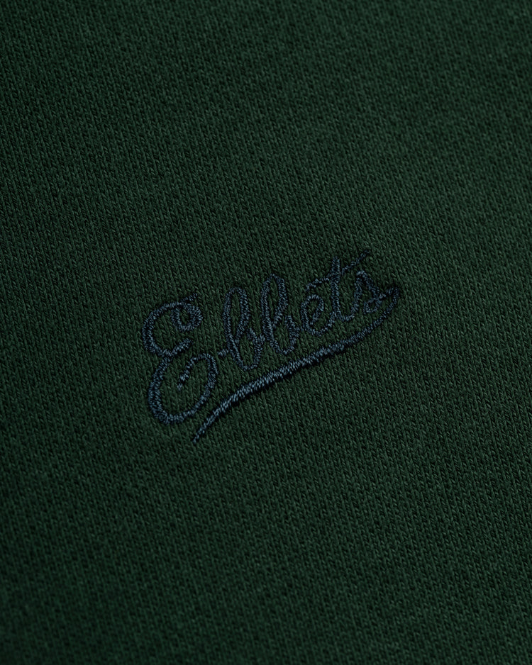 Close-up of the Ebbets Field Flannels Ebbets Chest Logo Heavyweight Cotton Terry Pullover Hoodie, featuring vintage-inspired dark green fabric and Ebbets embroidered in dark blue cursive on the chest.