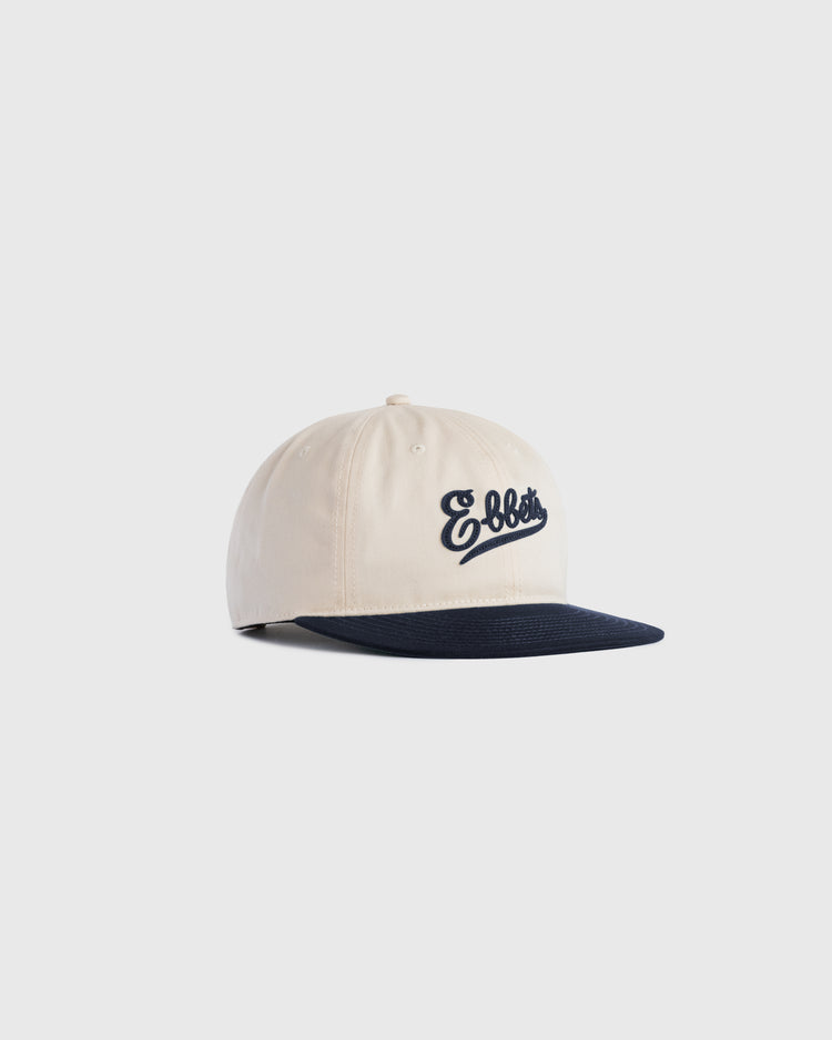 The Ebbets Field Flannels Ebbets Felt Script Cotton Twill Ballcap features beige cotton twill, a navy flat brim, and Ebbets embroidered in navy script on the front, displayed against a plain light background.