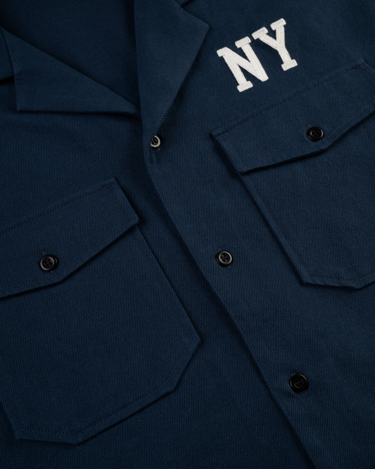 Close-up of the Ebbets Field Flannels Cotton Linen Grounds Crew Short Sleeve Workshirt in navy, featuring two front flap pockets, black buttons, and NY embroidered on the chest—a classic workwear style.