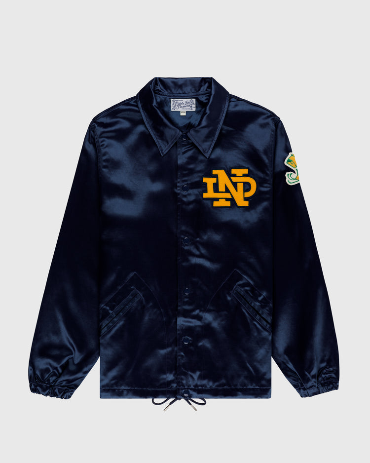 The EFF University of Notre Dame Satin Windbreaker Jacket is a shiny navy blue coach’s style featuring a yellow ND chest logo, colorful left sleeve patch, collar, snap buttons, two front pockets, and hem drawstrings.