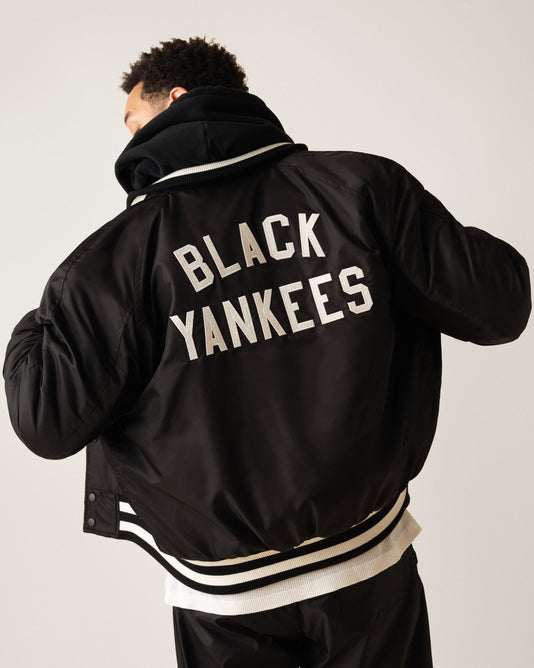 A person wearing a black varsity jacket with white stripes and the words BLACK YANKEES in large white letters on the back, shown from behind against a plain background.