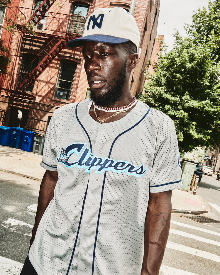 A man in an Ebbets Field Francisco Lindor Columbus Clippers Mesh Button-Up Jersey, a white New York cap, and a necklace stands on a city street with brick buildings and trees in the background.