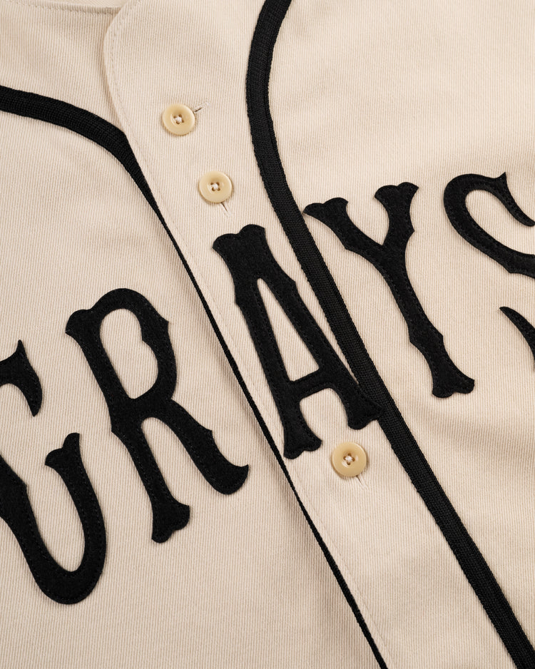 Close-up of the Ebbets Field Flannels Homestead Grays Cotton Twill Baseball Jersey, cream-colored with black lettering and trim, beige buttons, and GRAYS on the front—a tribute to the legendary Negro Leagues team.