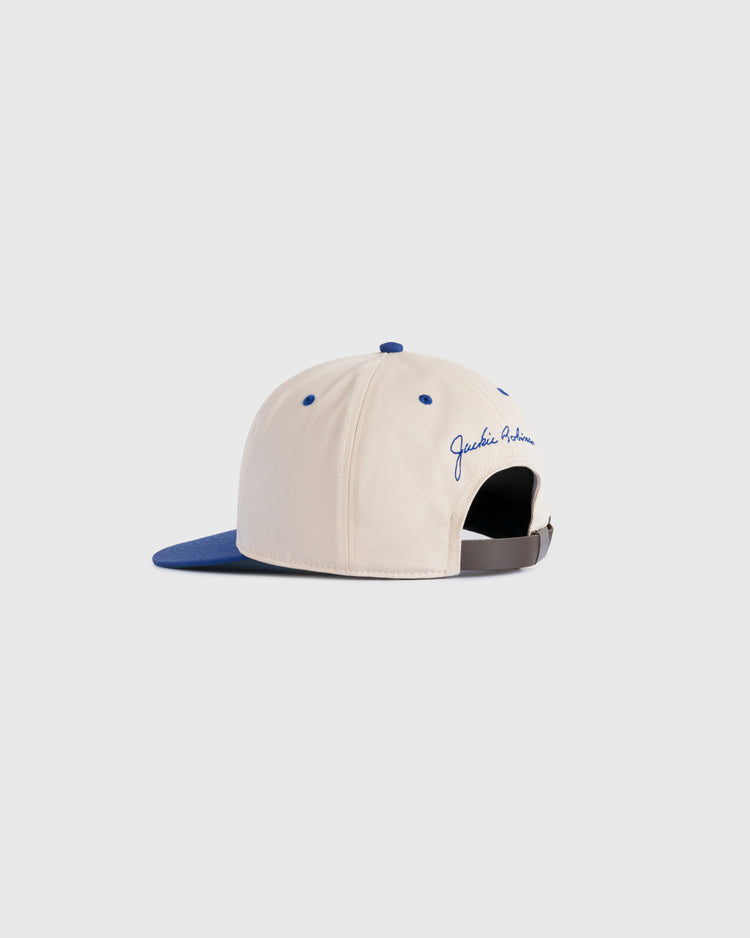 The Ebbets Field Jackie Robinson Day ‘42’ Cotton Twill Ballcap features a beige crown, blue brim, and black adjustable strap. Jackie Robinson is embroidered in blue script above the strap, shown on a light gray background.