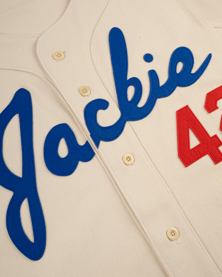 The Ebbets Field Jackie Robinson Cotton Twill Baseball Jersey features a cream color, blue “Jackie” script, red number 42, and white front buttons—an iconic tribute to the legendary player.