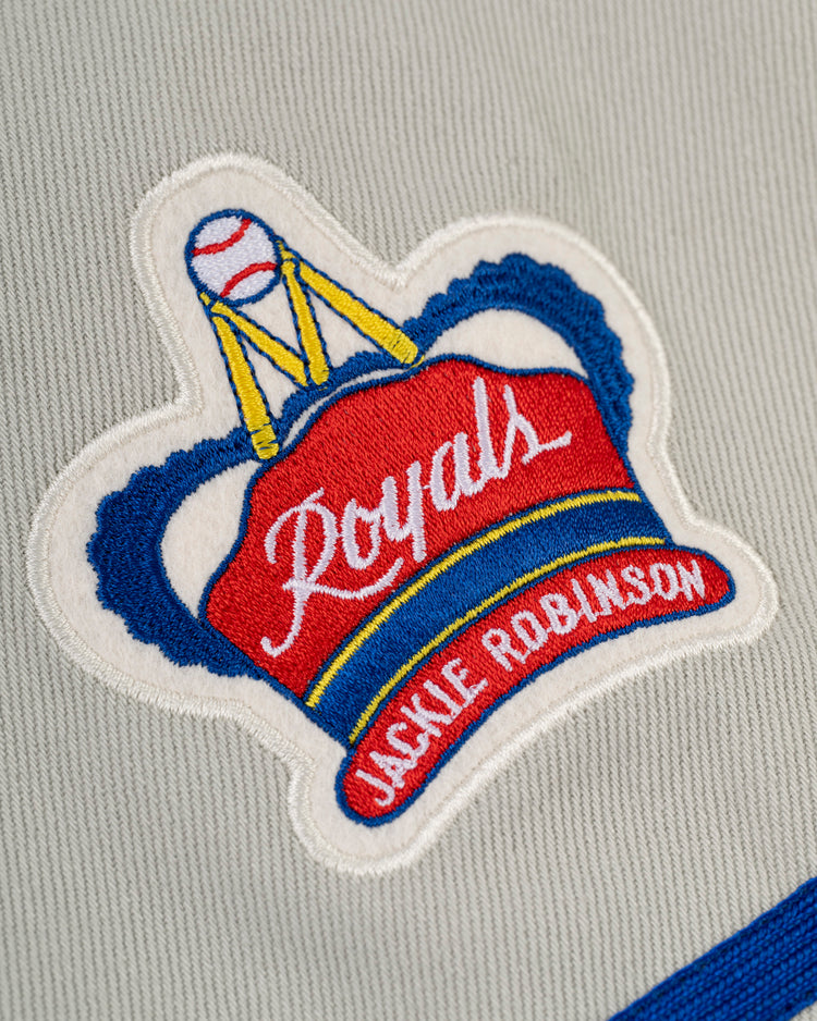 An embroidered patch on gray fabric inspired by the Montreal Royals Cotton Twill Baseball Jersey by Ebbets Field features a red and blue crown with Royals, a baseball on top, and Jackie Robinson in white on a yellow and blue band at the bottom.