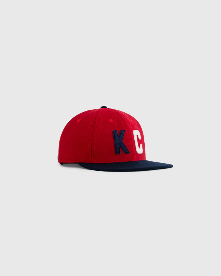 The EFF Kansas City Monarchs 1954 Authentic Wool Ballcap is red with a navy brim and features KC in blue and white on the front, inspired by the classic Negro Leagues team, shown on a light gray background.