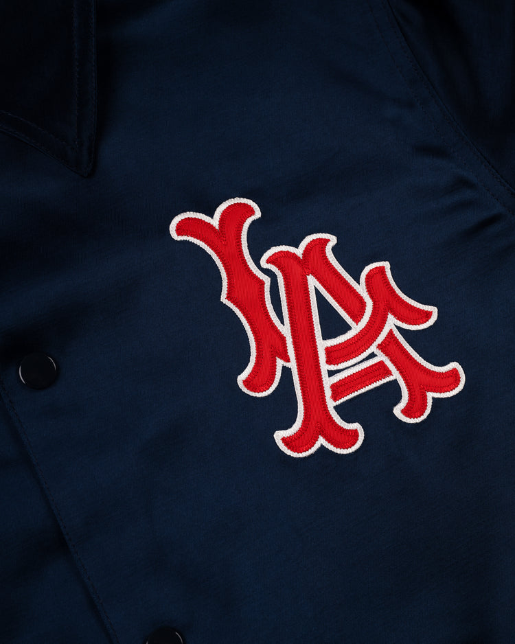 A close-up of the EFF Los Angeles Angels 1946 Satin Windbreaker shows a navy jacket with bold, red interlocking LA letters stitched on the front, white borders, and visible snaps—capturing vintage minor league baseball style.