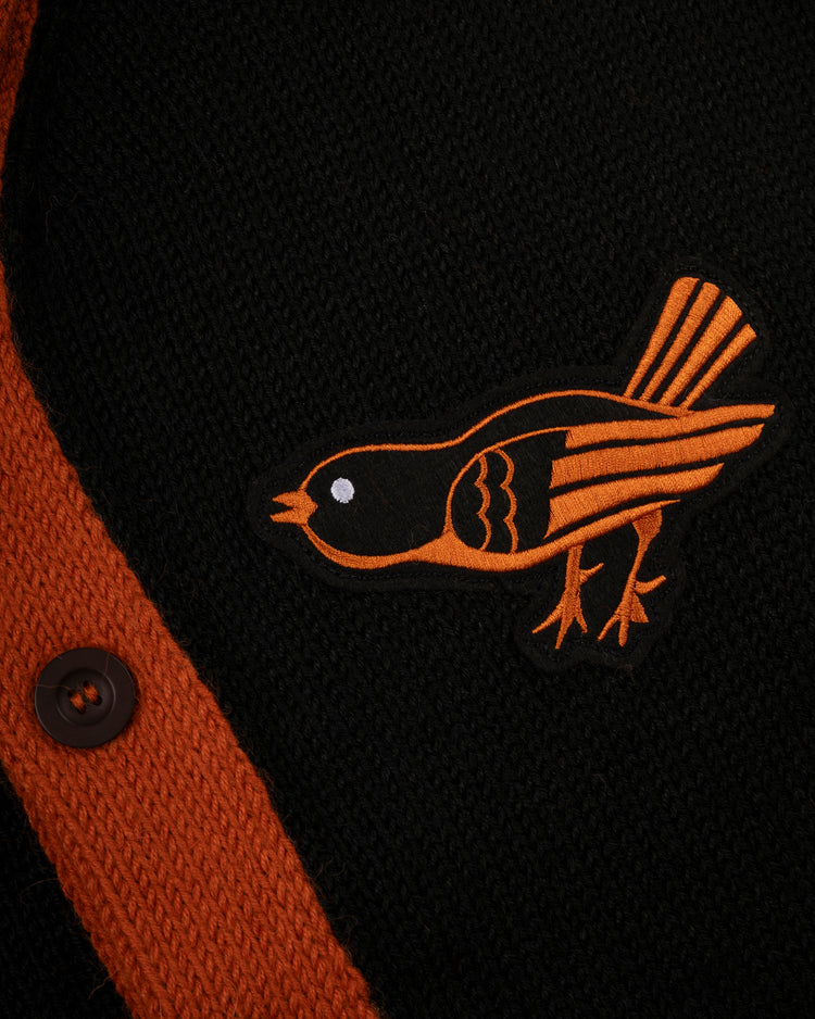 A black and orange embroidered bird patch is sewn onto the Baltimore Orioles 1964-65 Wool Shawl Collar Sweater by Ebbets Field Flannels, featuring black knit fabric with orange trim and a dark brown button on the left.