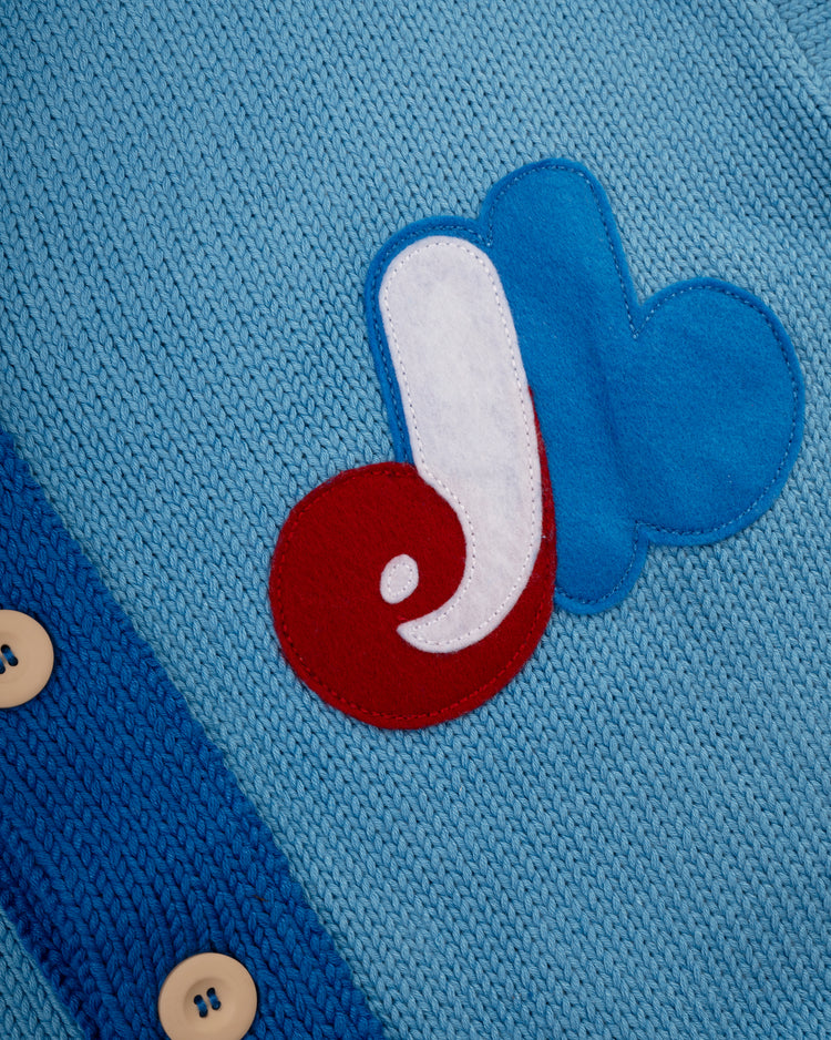 Close-up of light blue knit fabric with beige buttons and a patch featuring the Montreal Expos logo, on the Ebbets Field Flannels Montreal Expos 1969 Wool Shawl Collar Sweater, inspired by classic baseball styles.