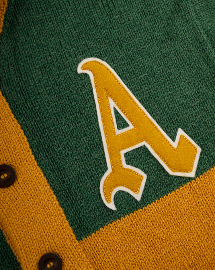 A close-up of the Ebbets Field Flannels Oakland Athletics 1969 Wool Shawl Collar Sweater, featuring green and yellow knit, black buttons, and a large yellow and white A patch inspired by vintage baseball sweaters.