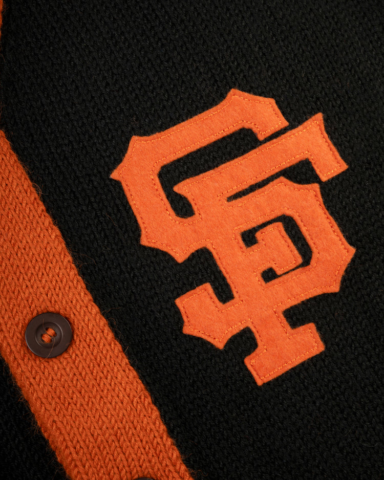 A close-up of the Ebbets Field Flannels San Francisco Giants 1958-1972 Wool Shawl Collar Sweater, featuring black knit fabric, an orange SF logo and trim, and a dark brown button—perfect for fans of vintage replica on-field sweaters.